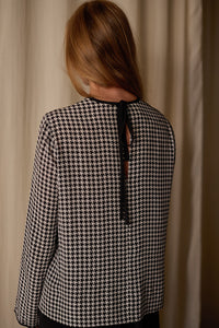 A person with long, light brown hair is standing facing away from the camera. They are wearing a black and white houndstooth patterned Qipao Blouse | Crepe de Chine Houndstooth, featuring a black ribbon tie at the neck. The background is a cream-colored curtain.