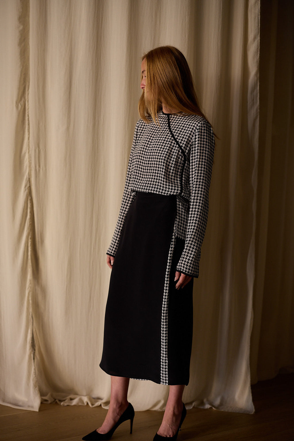 A person with long hair, dressed in a black and white houndstooth top paired with a Layered Skirt | Crepe de Chine featuring intricate houndstooth detailing along the side. They are wearing black high-heeled shoes and standing against a beige, softly draped fabric backdrop.