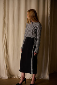 A person with long hair, dressed in a black and white houndstooth top paired with a Layered Skirt | Crepe de Chine featuring intricate houndstooth detailing along the side. They are wearing black high-heeled shoes and standing against a beige, softly draped fabric backdrop.