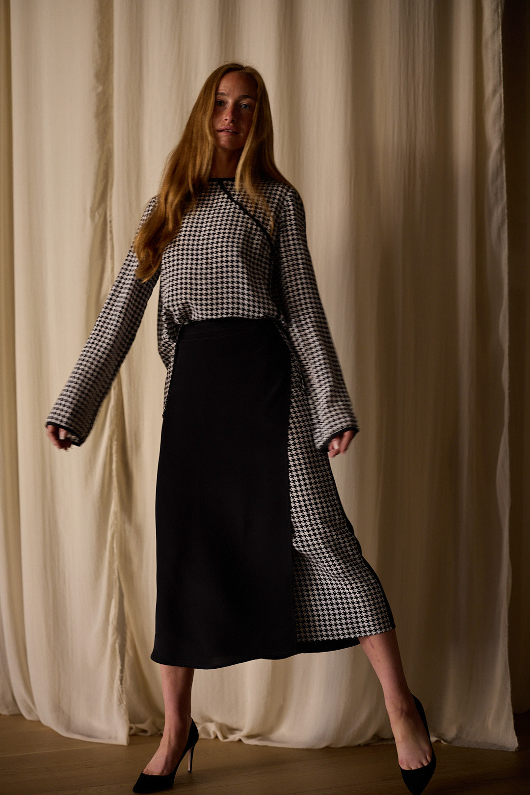 A woman with long red hair stands against a backdrop of light-colored draped fabric. She is wearing a long-sleeved black and white houndstooth top and a Layered Skirt | Crepe de Chine. Posing with one foot slightly raised, she showcases her elegant black high-heeled shoes.