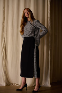 A woman with long brown hair is standing in front of a beige curtain. She is wearing a black and white Qipao Blouse | Crepe de Chine Houndstooth, a black skirt with a custom houndstooth side panel, and black high heels. She is posing with her hands on her hips and looking confidently at the camera.