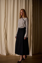 Load image into Gallery viewer, A woman with long blonde hair stands in front of light cream-colored drapes. She is wearing a handmade, long-sleeved Qipao Blouse | Crepe de Chine Custom Houndstooth paired with a high-waisted black pleated skirt and black high heels. She looks directly at the camera with a neutral expression.