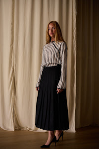 A woman with long blonde hair stands in front of light cream-colored drapes. She is wearing a handmade, long-sleeved Qipao Blouse | Crepe de Chine Custom Houndstooth paired with a high-waisted black pleated skirt and black high heels. She looks directly at the camera with a neutral expression.