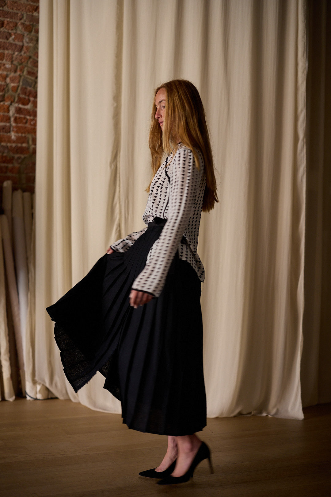 A woman with long red hair, wearing a Qipao Blouse | Crepe de Chine Custom Houndstooth and a flowing black skirt, stands sideways, holding her skirt with one hand. She is in a room with wooden floors, a brick wall, and cream-colored curtains in the background.