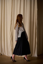 Load image into Gallery viewer, A woman with long, reddish hair is standing sideways against a backdrop of cream-colored curtains. She is wearing a Qipao Blouse | Crepe de Chine Custom Houndstooth with long sleeves, paired with a long, black pleated skirt. She has black high-heeled shoes on and her left arm is slightly bent back.