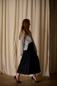 A woman with long, reddish hair is standing sideways against a backdrop of cream-colored curtains. She is wearing a Qipao Blouse | Crepe de Chine Custom Houndstooth with long sleeves, paired with a long, black pleated skirt. She has black high-heeled shoes on and her left arm is slightly bent back.