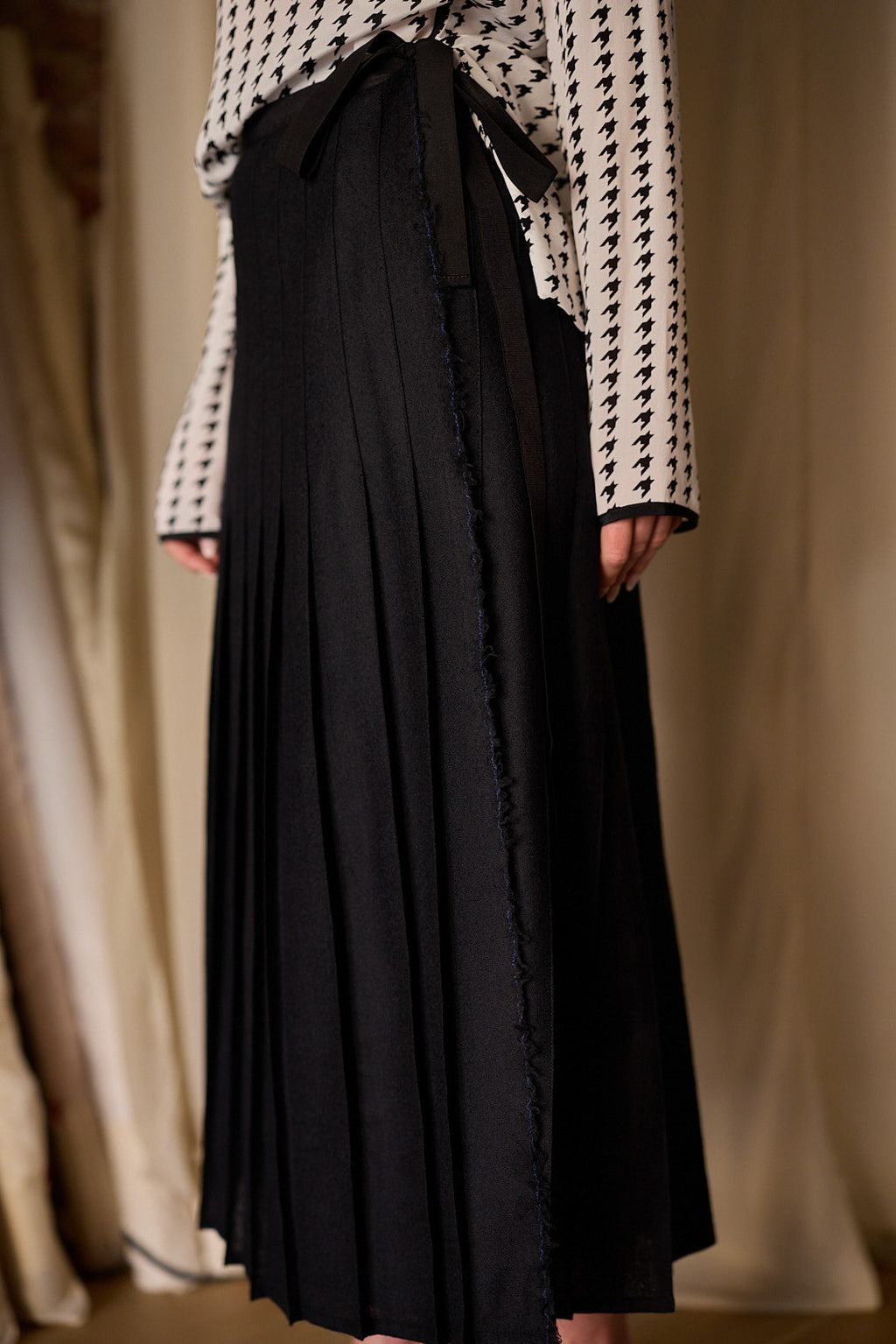 Wearing the Hand Pleated Wrap Skirt crafted from Japanese wool gauze, a person pairs it with a long-sleeved top showcasing a black and white houndstooth pattern. The image highlights the outfit's details, emphasizing its textures and patterns while adhering to zero-waste design principles.