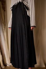 Load image into Gallery viewer, A person is wearing a black, Hand Pleated Wrap Skirt - Japanese Wool Gauze paired with a white, long-sleeved blouse featuring a black houndstooth pattern. The details and texture of the clothing, made from Japanese wool gauze, are highlighted in this side profile shot.