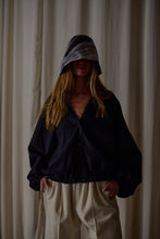 Load image into Gallery viewer, A person stands in front of a light-colored curtain, wearing a loose-fitting dark jacket and light-colored pants. They have long hair and are partially obscured by a Silk Organza Ribbon Cloche | Black, Reversible that casts a shadow over their face. Their hands are casually placed in their pockets.
