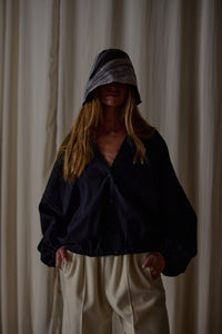 A person stands in front of a light-colored curtain, wearing a loose-fitting dark jacket and light-colored pants. They have long hair and are partially obscured by a Silk Organza Ribbon Cloche | Black, Reversible that casts a shadow over their face. Their hands are casually placed in their pockets.