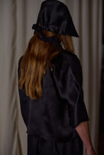 Load image into Gallery viewer, A person with long brown hair is seen from the back, wearing a Silk Organza Ribbon Cloche | Black, Reversible tied under the chin and a matching black satin top. The background is out of focus with light-colored curtains.