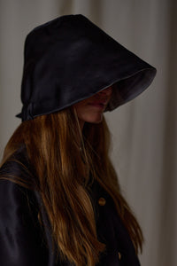 A person with long, brown hair is wearing a dark hooded garment that partially obscures their face, giving them an air of mystery. The background is a neutral-colored curtain, adding to the subtle yet intriguing scene. The overall aesthetic could be enhanced with a Silk Organza Ribbon Cloche | Black, Reversible to complete the look.