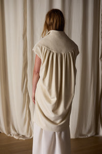 A person with long hair, seen from behind, is wearing a loose, cream-colored sleeveless top with a wide collar that drapes down the back. The Poet Vest Coat | Undyed, handmade in-house from cashmere, complements their look. They stand before off-white curtains on a wooden floor.