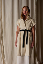 Load image into Gallery viewer, A woman with long, light brown hair is standing in front of beige curtains. She is wearing a beige Poet Vest Coat | Undyed, handmade in-house, with a high collar and a black belt tied around the waist. The coat, resembling an elegant cashmere vest, reaches her knees, and she has a calm expression on her face.
