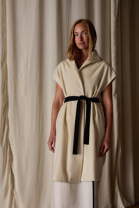 A woman with long, light brown hair is standing in front of beige curtains. She is wearing a beige Poet Vest Coat | Undyed, handmade in-house, with a high collar and a black belt tied around the waist. The coat, resembling an elegant cashmere vest, reaches her knees, and she has a calm expression on her face.
