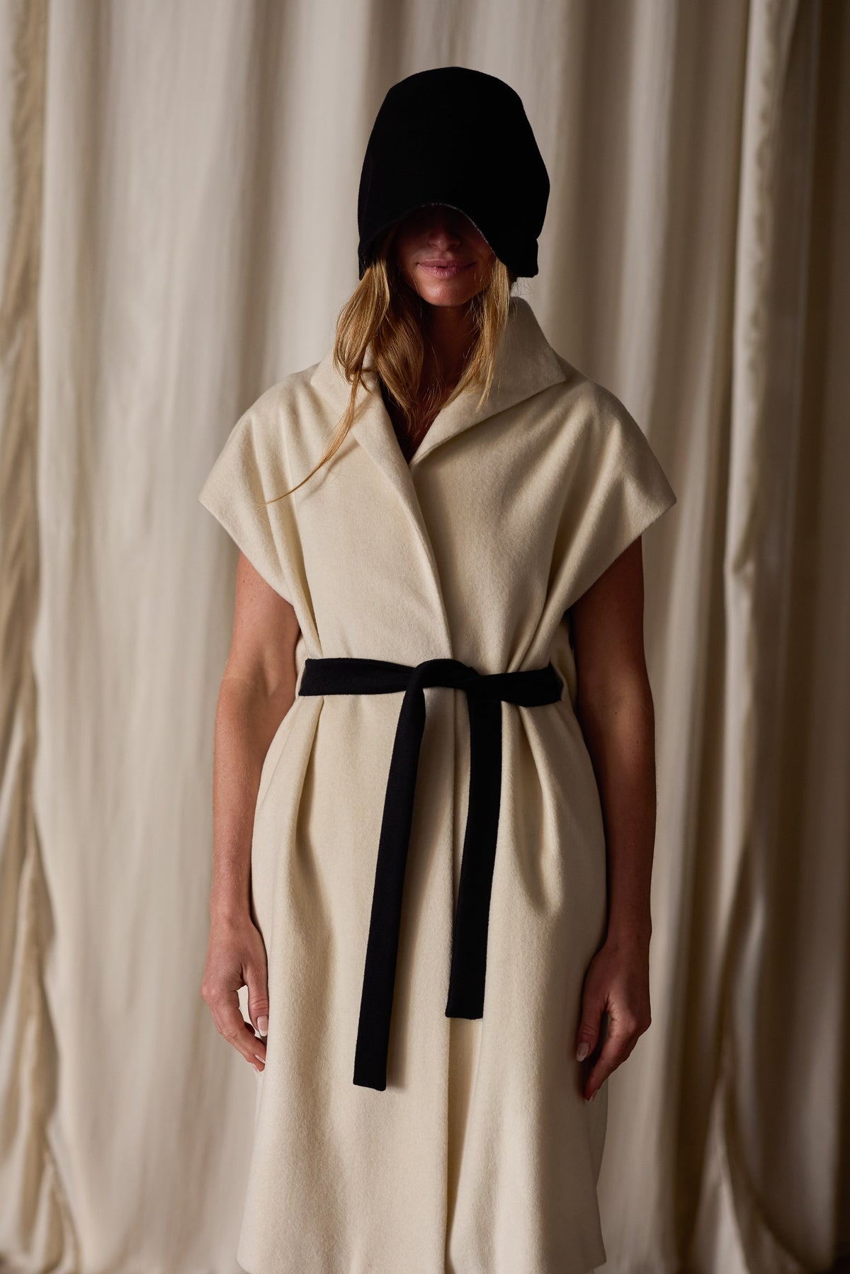 A person stands in front of a neutral backdrop, wearing a sleeveless Poet Vest Coat | Undyed with a wide collar. Handmade in-house, the cream-colored piece is cinched at the waist with a black belt. They also wear a black head covering that obscures their face, with long hair visible underneath.