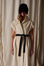 Load image into Gallery viewer, A person stands in front of a neutral backdrop, wearing a sleeveless Poet Vest Coat | Undyed with a wide collar. Handmade in-house, the cream-colored piece is cinched at the waist with a black belt. They also wear a black head covering that obscures their face, with long hair visible underneath.