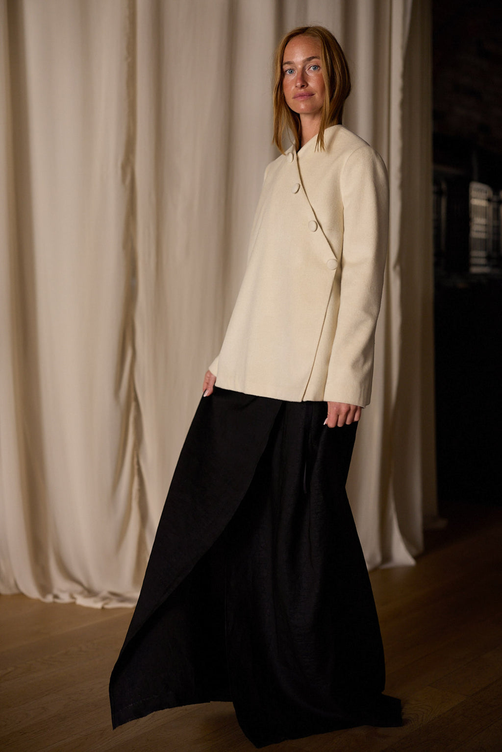 A person with long, straight hair stands indoors against a backdrop of neutral curtains. They are wearing a Qipao Jacket | Cashmere Undyed and a long, flowing, dark-colored skirt. They are smiling slightly and gazing toward the camera.