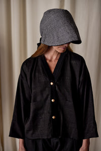 A person is wearing a dark, loose-fitting jacket with three light-colored buttons down the front and large pockets. They have on a Cashmere Ribbon Cloche | Black, Reversible with a wide brim covering their face. The background is blurred with soft, light-colored curtains.