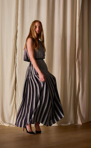 A person with long hair is wearing the Hand Pleated Column Dress Reversible | Custom Houndstooth Stripe made from 100% silk, featuring a pattern of vertical black and white stripes. They are standing on a wooden floor in front of light-colored, textured drapes and are wearing black high-heeled shoes.