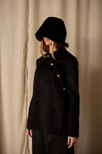 Load image into Gallery viewer, A person stands against a beige curtain, wearing a black wide-brimmed hat that obscures their face. They are dressed in a Qipao Jacket | Cashmere Black with gold buttons and matching black trousers.