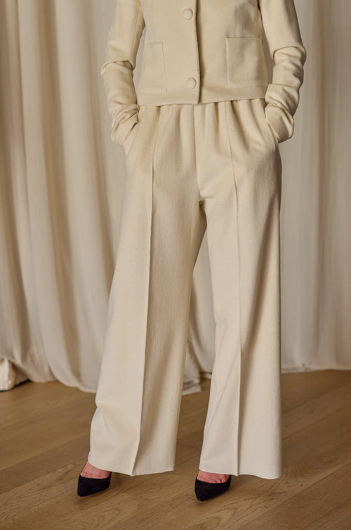 A person stands with their hands in the pockets of Cashmere Trousers l Undyed. They are also wearing a matching cream-colored jacket from Atelier Lan Jaenicke with long sleeves and black heels. The background features beige curtains and a wooden floor. The shot is from the shoulders down.