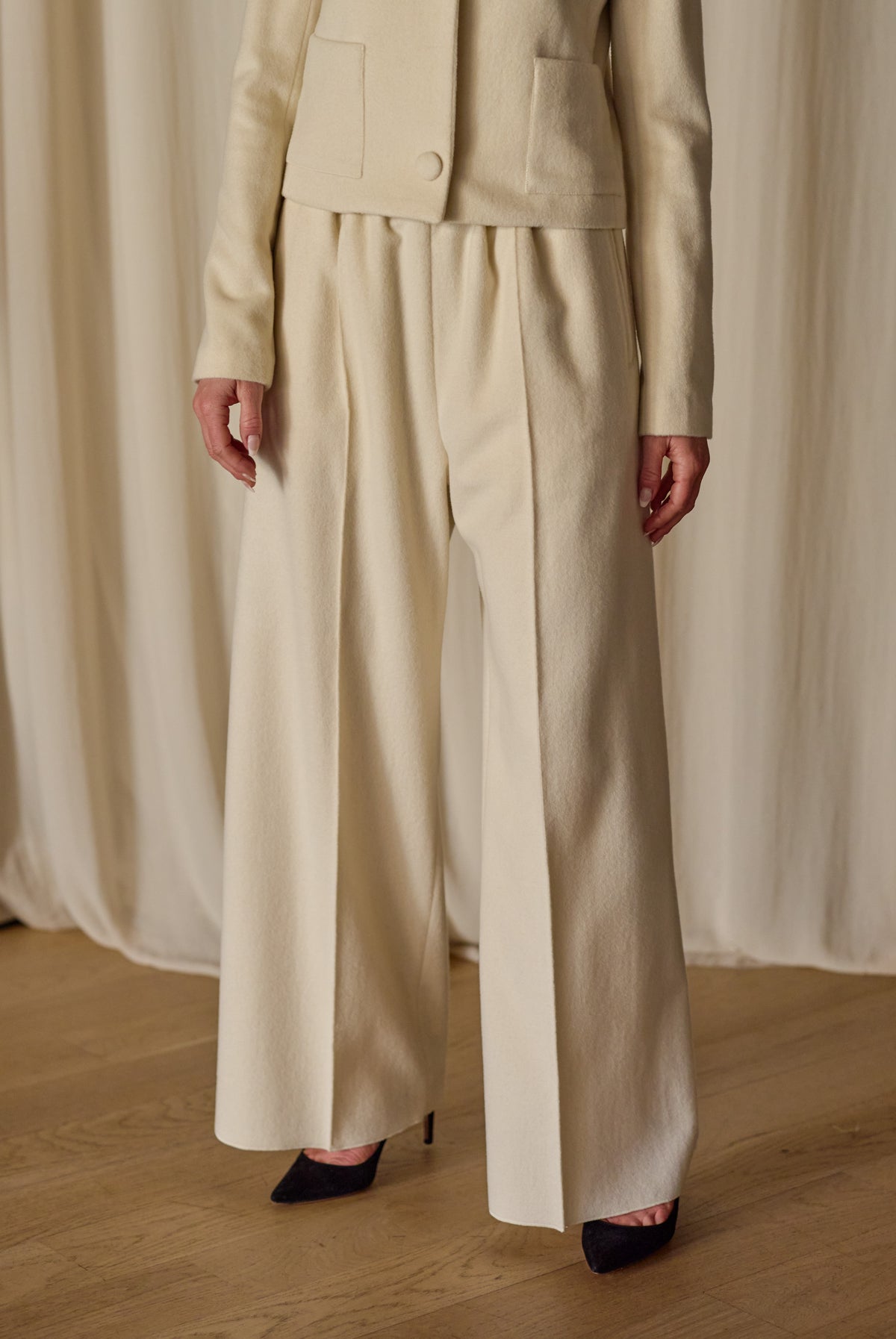 A person stands in a light-colored room wearing a cream-colored outfit by Atelier Lan Jaenicke. The attire consists of a long-sleeve jacket and Cashmere Trousers l Undyed. Only the body from the shoulders down is visible, and the person is wearing black high-heeled shoes.