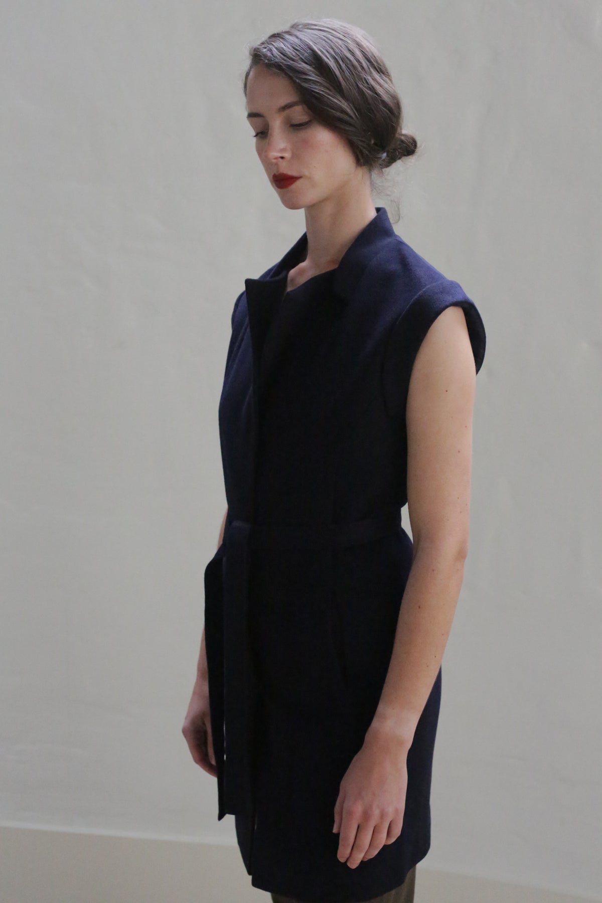A person with dark hair tied back is standing with their eyes closed, wearing a sleeveless Lexington | Navy vest-coat with a belt. The background is plain and light-colored, creating a minimalist and serene atmosphere.