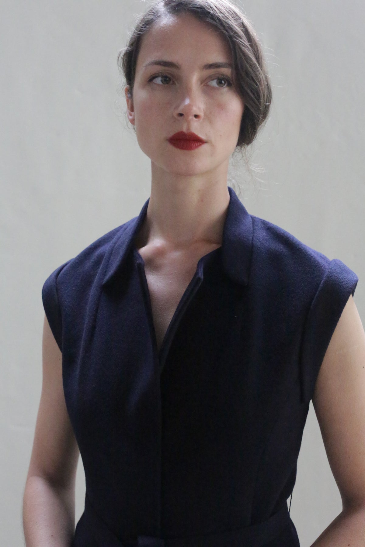 A person with long hair styled in a side braid is wearing the Lexington | Navy, a dark sleeveless, collared top that resembles a vest-coat. They have a serious expression, red lipstick, and are looking slightly to the side against a plain light background.