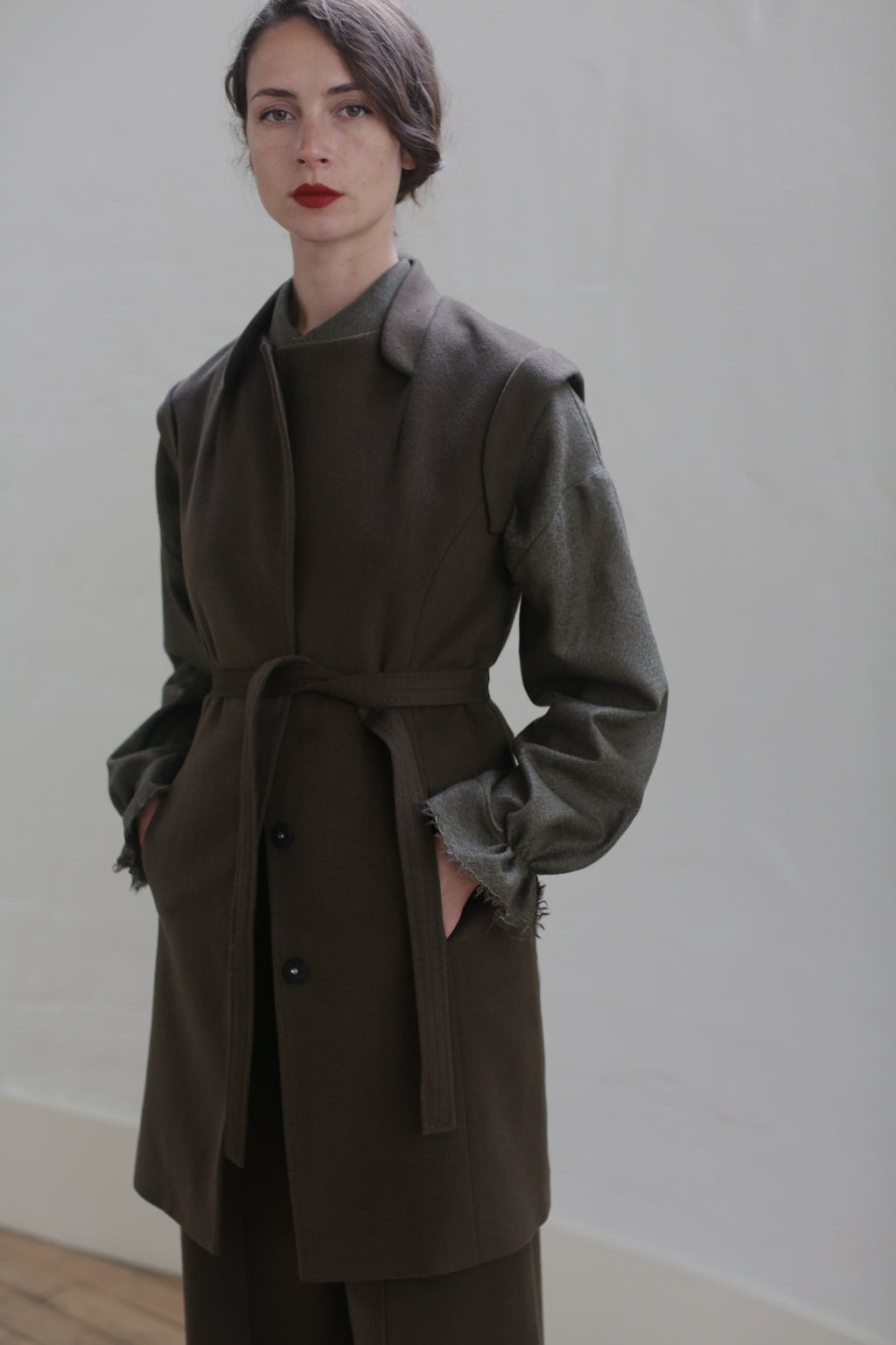 A woman with red lipstick is standing against a light-colored wall. She is wearing a long, dark brown vest-coat with a belted waistline from Lexington | Olive over a frayed green shirt. Her hands are in the coat pockets, and her dark hair is styled in a low updo.