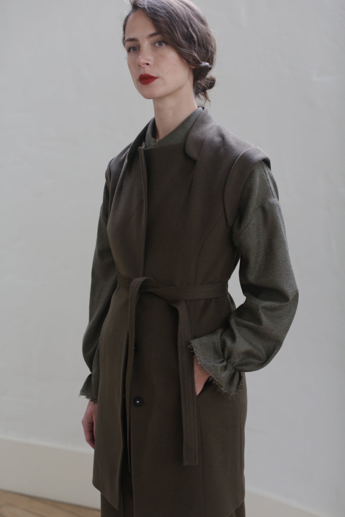 A person with long, dark hair stands against a plain light-colored wall. They are wearing the Lexington | Olive with a belted waistline over a green, long-sleeved top and have a neutral expression with bold red lips, looking slightly to the side.