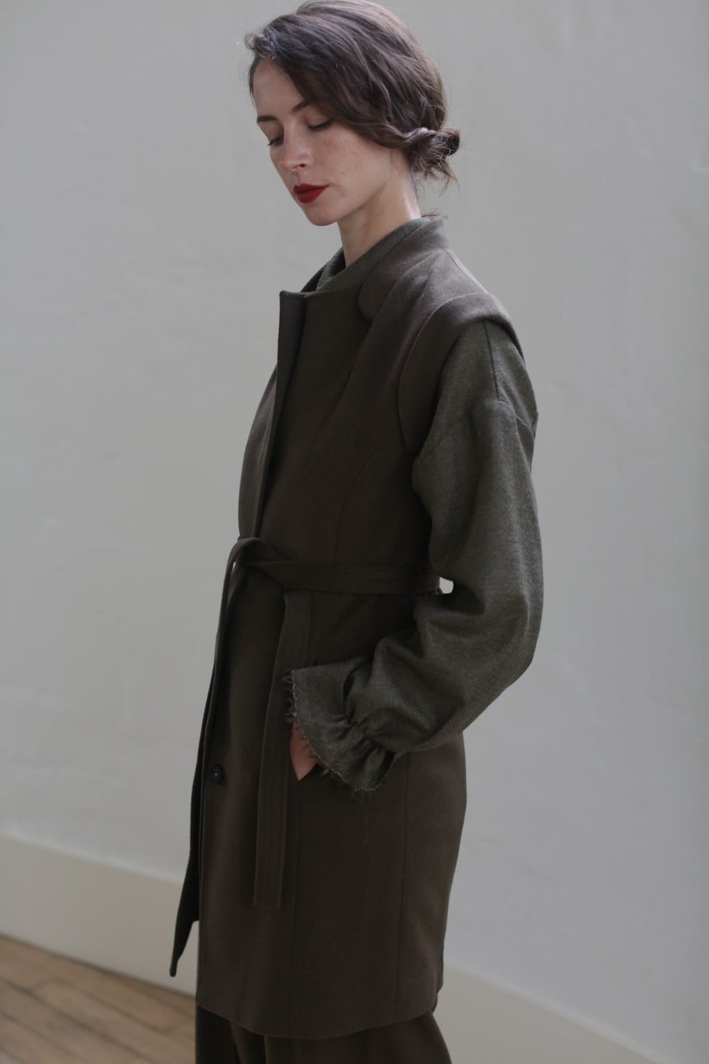 A person with short dark hair is standing against a plain light-colored wall. They are wearing a Lexington | Olive, featuring a belted waistline over a long-sleeved shirt. The person's hands are in their coat pockets, and they appear to be looking down, with a solemn expression.