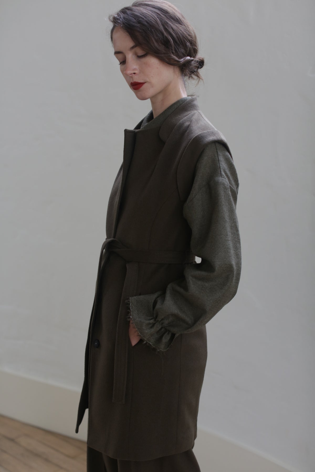 A person with short dark hair is standing against a plain light-colored wall. They are wearing a Lexington | Olive, featuring a belted waistline over a long-sleeved shirt. The person's hands are in their coat pockets, and they appear to be looking down, with a solemn expression.
