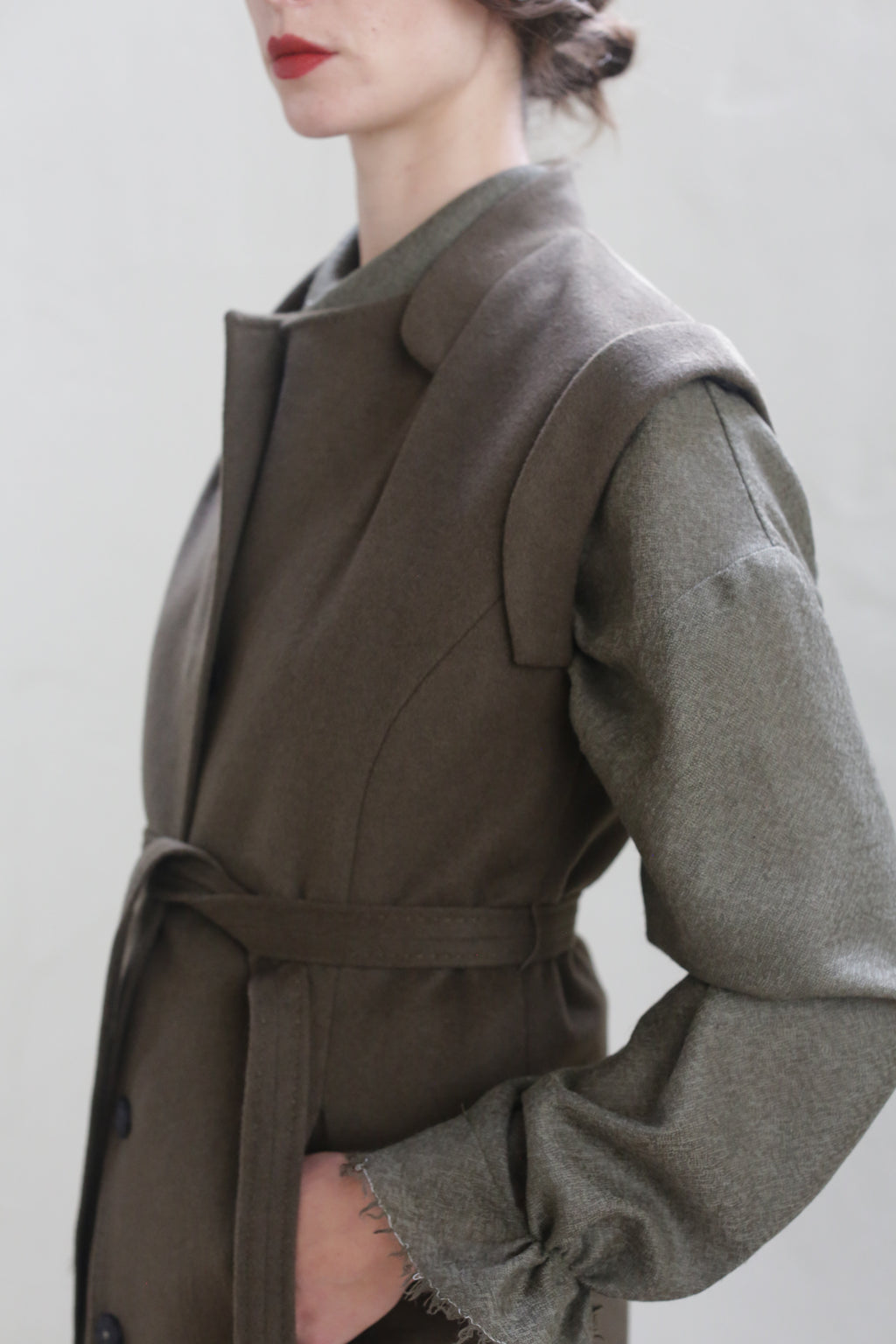 A person with red lipstick is partially seen from the side, wearing a Lexington | Olive with a belted waistline over a gray long-sleeve top. The person's face is not fully visible. The background is plain and gray.