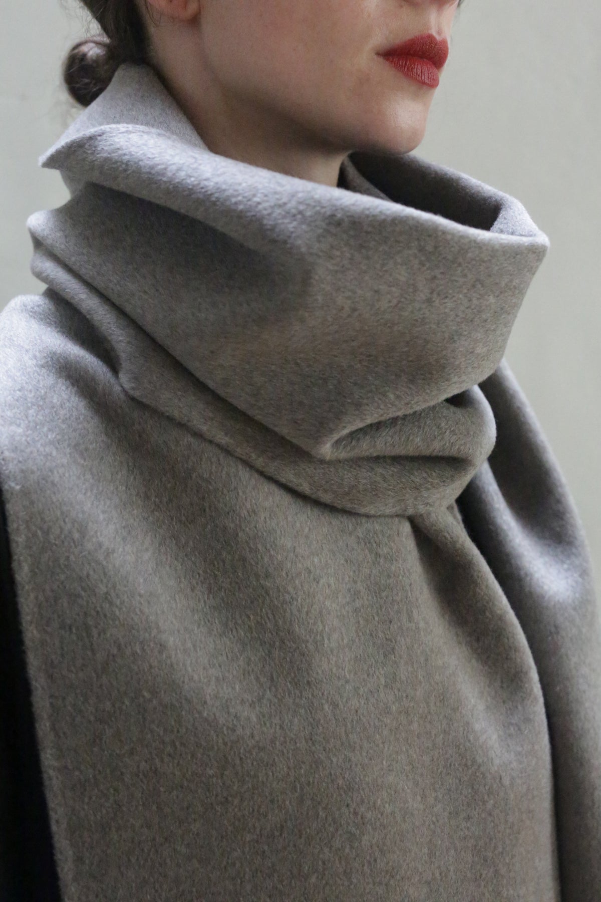 A close-up of a person wearing a Long Scarf Double-face Cashmere | Undyed Natural that wraps around their neck and shoulders. The handmade scarf appears thick and cozy, partially covering the person's mouth. They have fair skin and are wearing red lipstick.