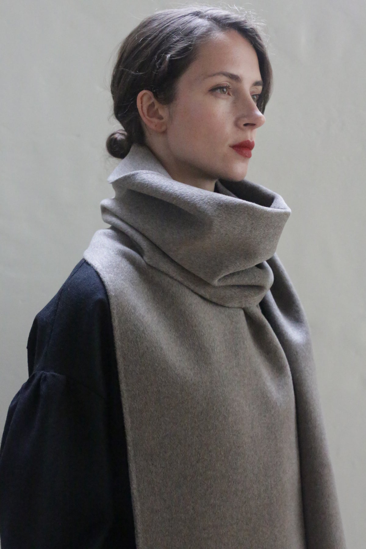 A person with fair skin and brown hair is looking to the side, wearing a Long Scarf Double-face Cashmere | Undyed Natural around their neck. They are dressed in a black top, with hair styled in a low bun. The background is plain and neutral in color, highlighting the elegance of their handmade accessory.