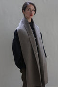 A person with straight dark hair is wearing a dark long-sleeve shirt, brown pants, and a Long Scarf Double-face Cashmere | Undyed Natural draped over their shoulders. They are standing against a plain, light-colored background, looking slightly to the side.