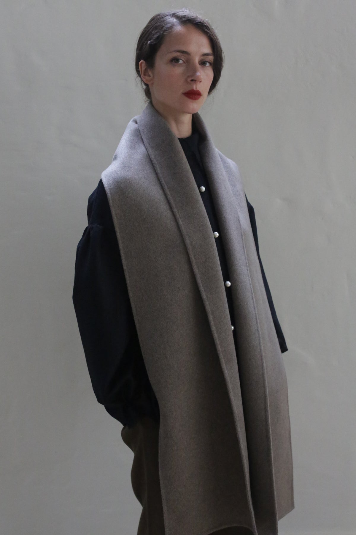 A person with straight dark hair is wearing a dark long-sleeve shirt, brown pants, and a Long Scarf Double-face Cashmere | Undyed Natural draped over their shoulders. They are standing against a plain, light-colored background, looking slightly to the side.