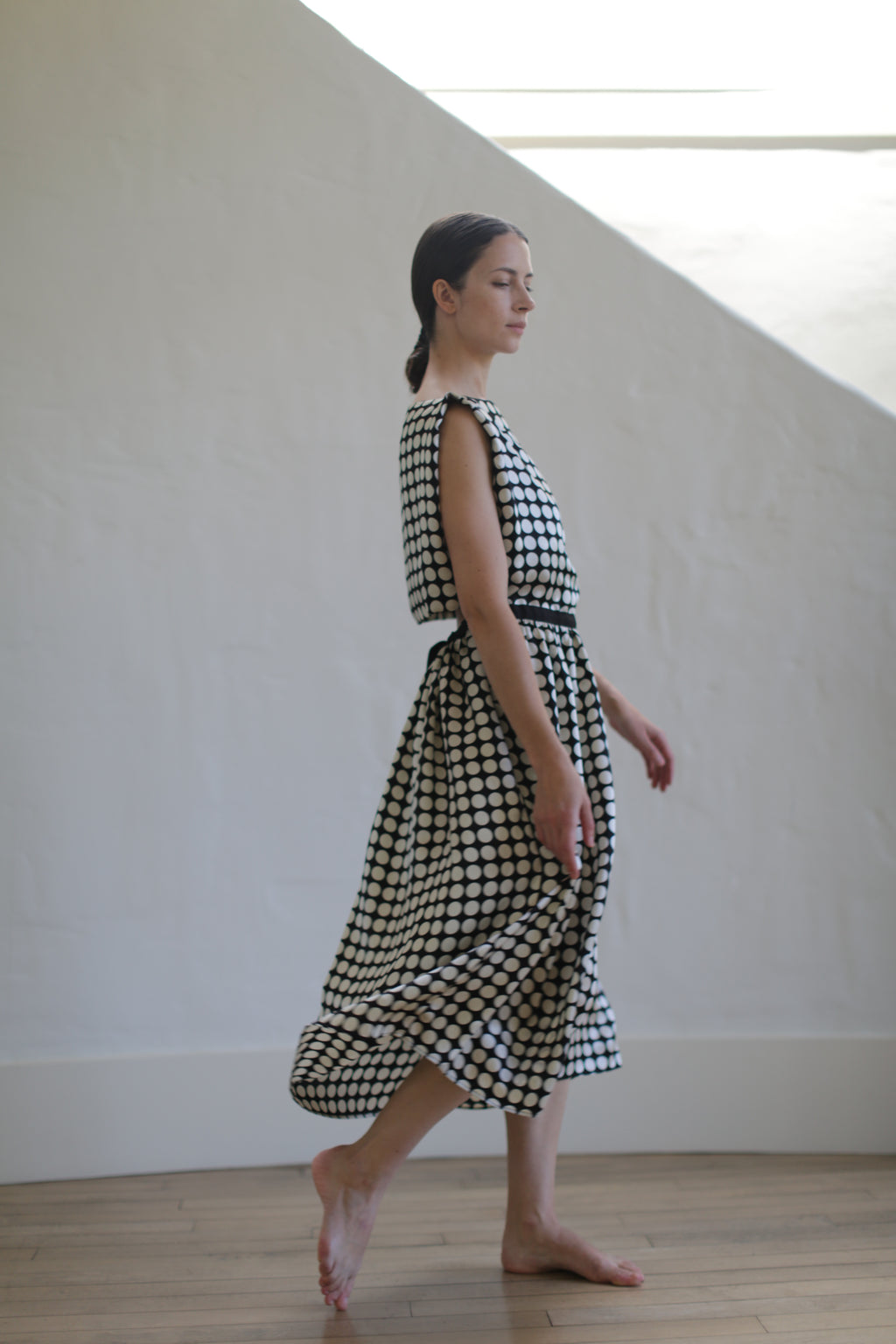 A woman with dark hair tied back is walking barefoot indoors. She is wearing a sleeveless, ankle-length Magnolia Dress | W/B Polkadot, made from luxurious silk charmeuse. The open back design accentuates the elegance of the dress. A light-colored wall in the background creates a calm and minimalist setting.

