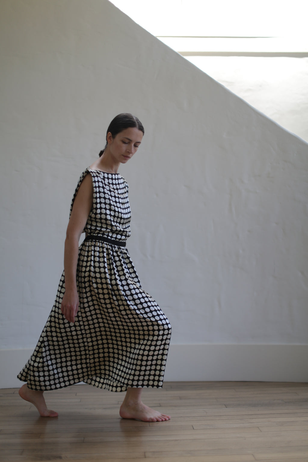 A woman with long dark hair is walking barefoot on a wooden floor. She is wearing a sleeveless, Magnolia Dress | W/B Polkadot with an open back design that flows around her legs. The background is a minimalist white wall with natural light filtering in from an unseen window above.