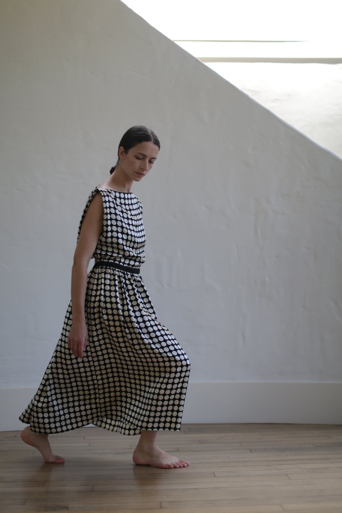 A woman with long dark hair is walking barefoot on a wooden floor. She is wearing a sleeveless, Magnolia Dress | W/B Polkadot with an open back design that flows around her legs. The background is a minimalist white wall with natural light filtering in from an unseen window above.