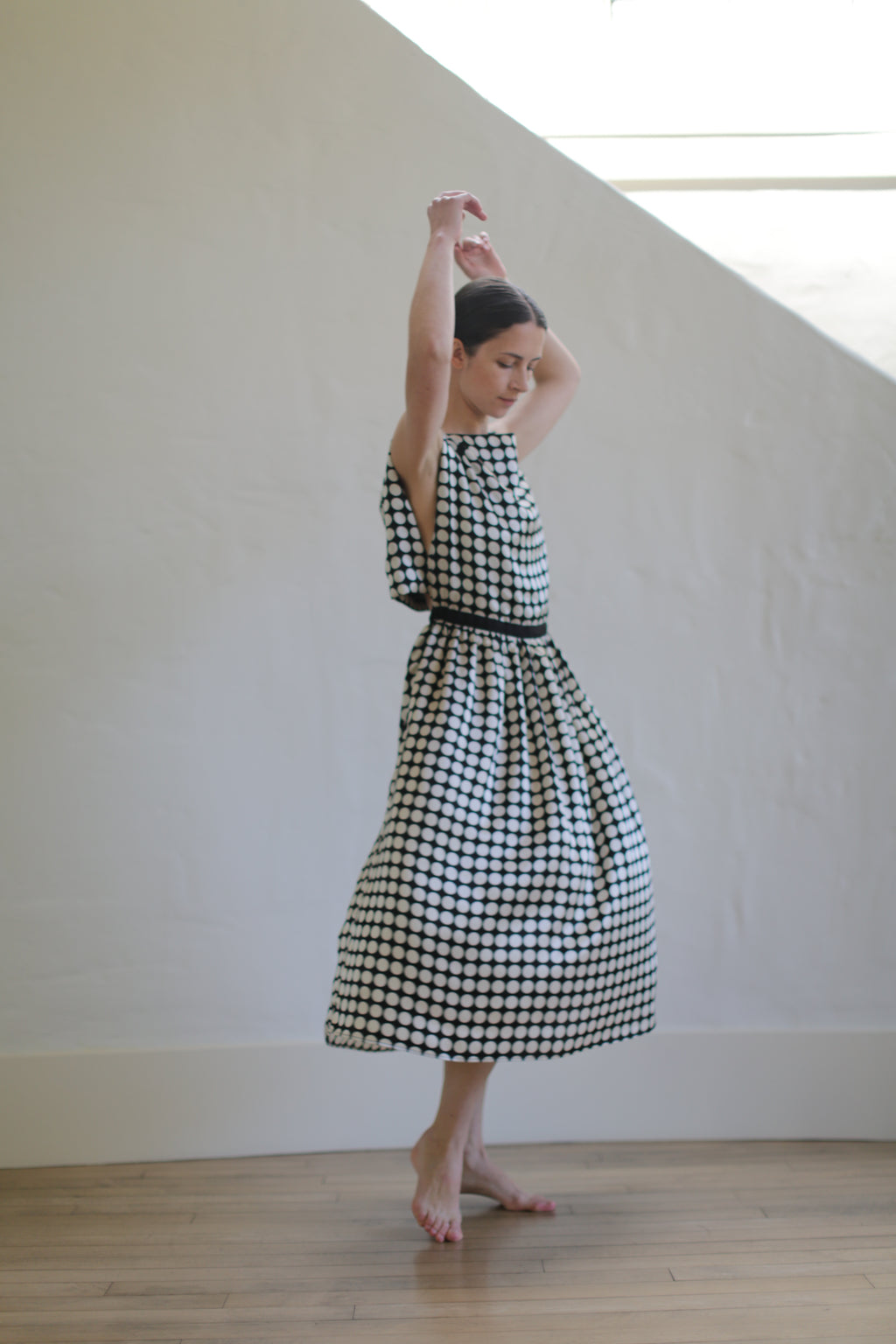 A woman in a sleeveless Magnolia Dress | W/B Polkadot with an open back design is standing against a light-colored wall, her arms raised gracefully above her head. She appears to be posing or preparing for a dance, with one foot slightly raised off the ground.