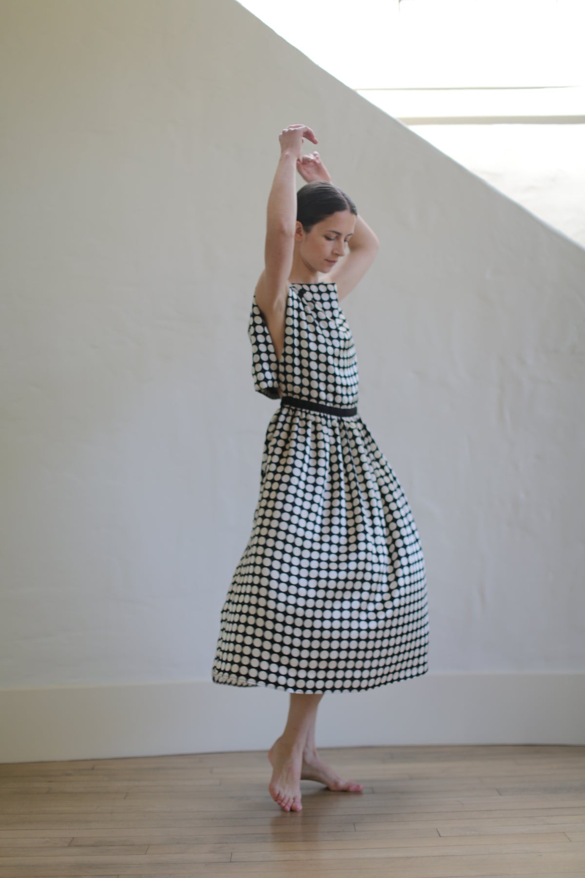 A woman in a sleeveless Magnolia Dress | W/B Polkadot with an open back design is standing against a light-colored wall, her arms raised gracefully above her head. She appears to be posing or preparing for a dance, with one foot slightly raised off the ground.
