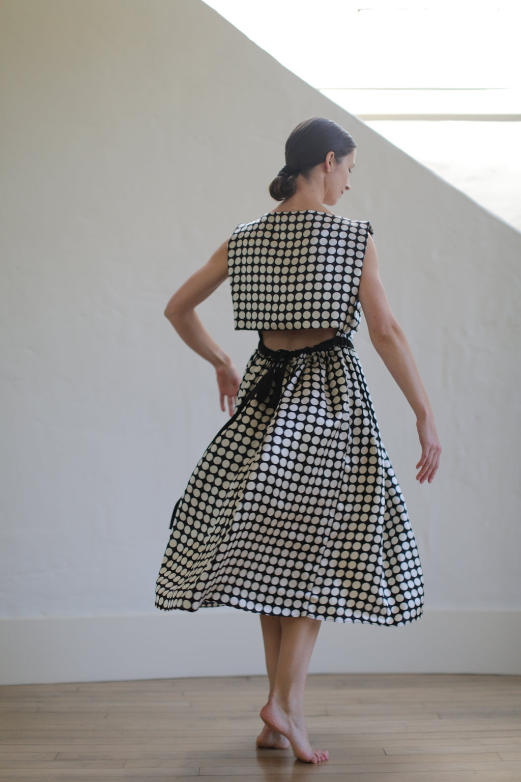 A woman with dark hair is dancing barefoot in a spacious, brightly lit room. She is wearing the Magnolia Dress | W/B Polkadot, which features an open back design, cropped top, and high-waisted skirt, creating an elegant, vintage look.