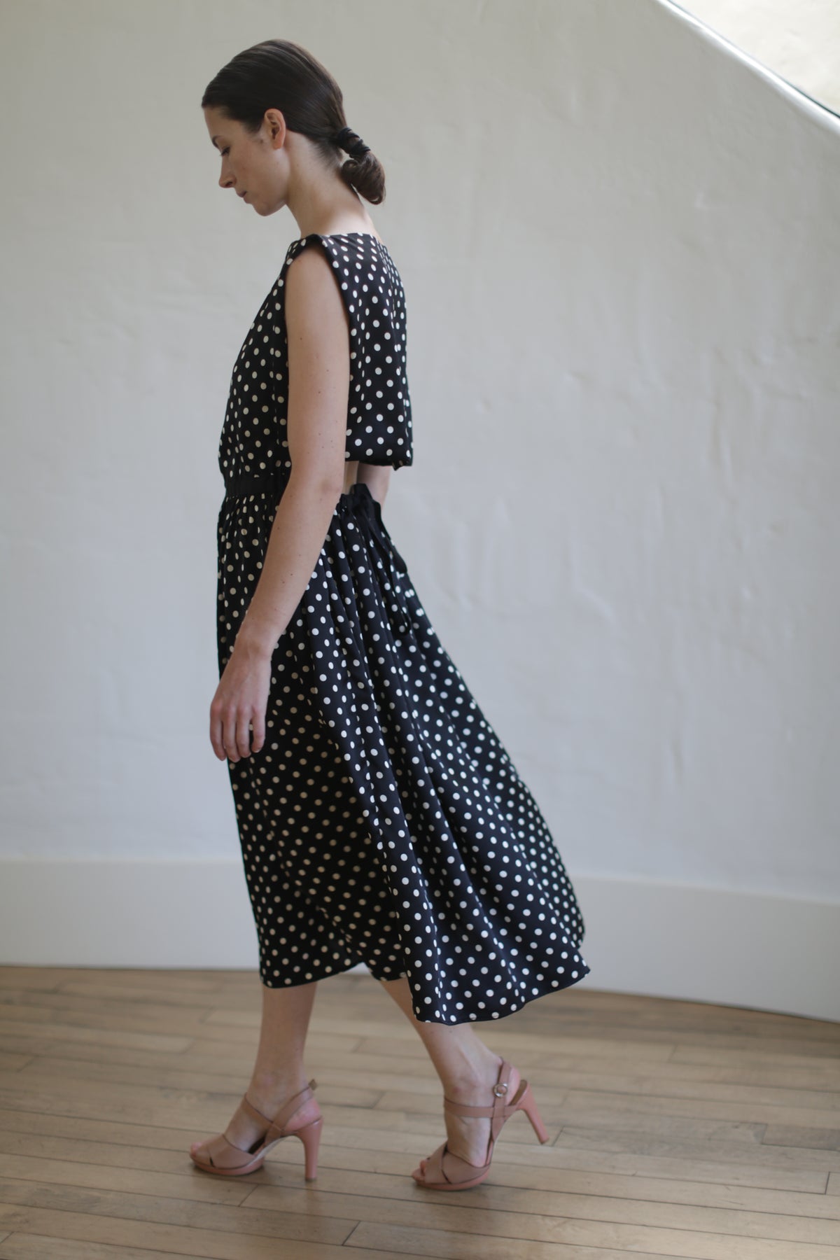 A person with dark hair tied back in a low bun is walking indoors wearing a sleeveless Magnolia Dress | B/W Polkadot with pink heeled sandals. The dress has a high slit and an open back, subtly highlighting the handmade design. The background is a plain, light-colored wall with a wooden floor.