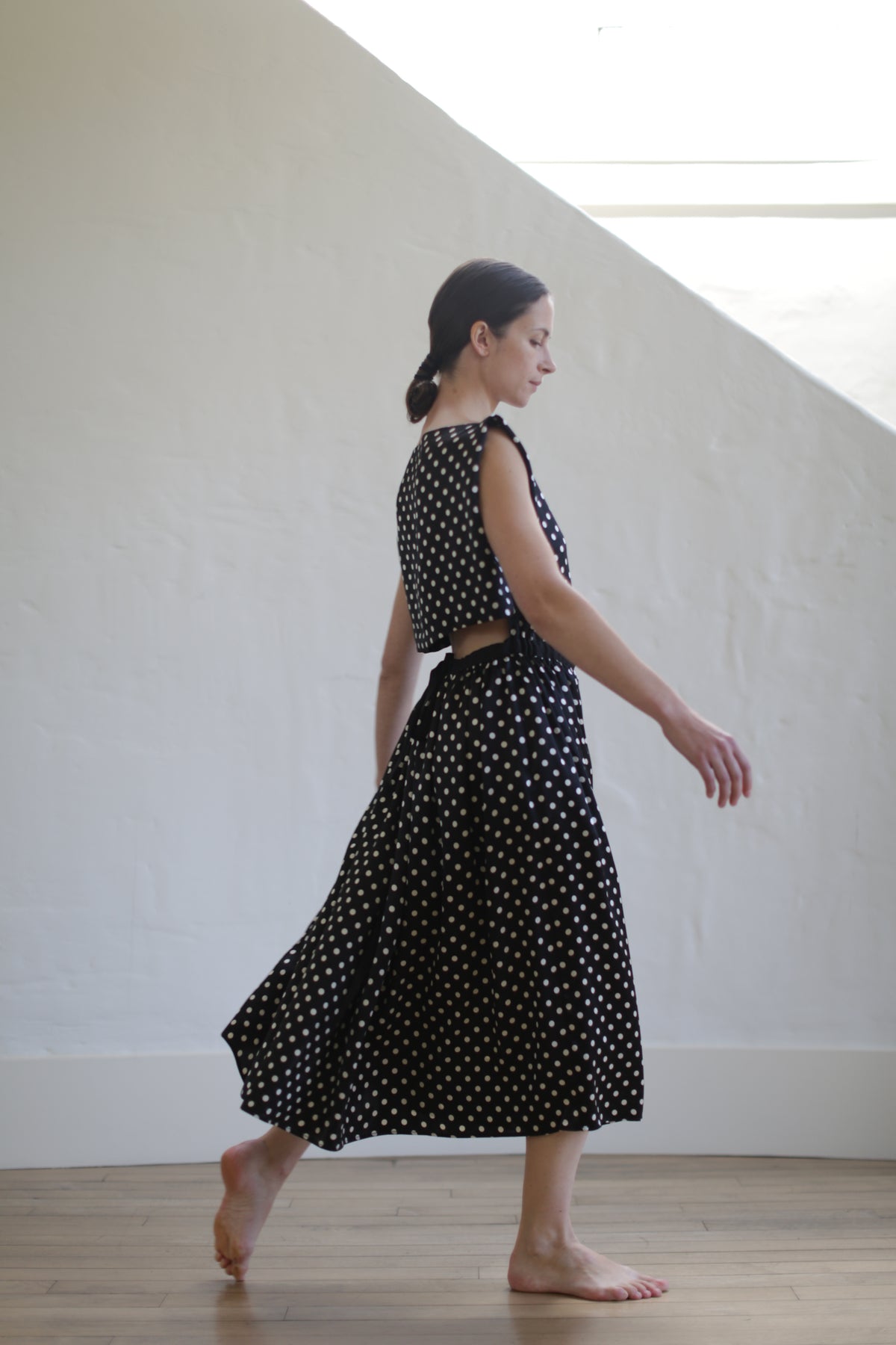 A woman with dark hair, tied back, wearing a Magnolia Dress | B/W Polkadot. She is barefoot, walking indoors on a wooden floor, with a light-filled, white, angled-wall background. Her outfit includes an elegant open-back design.