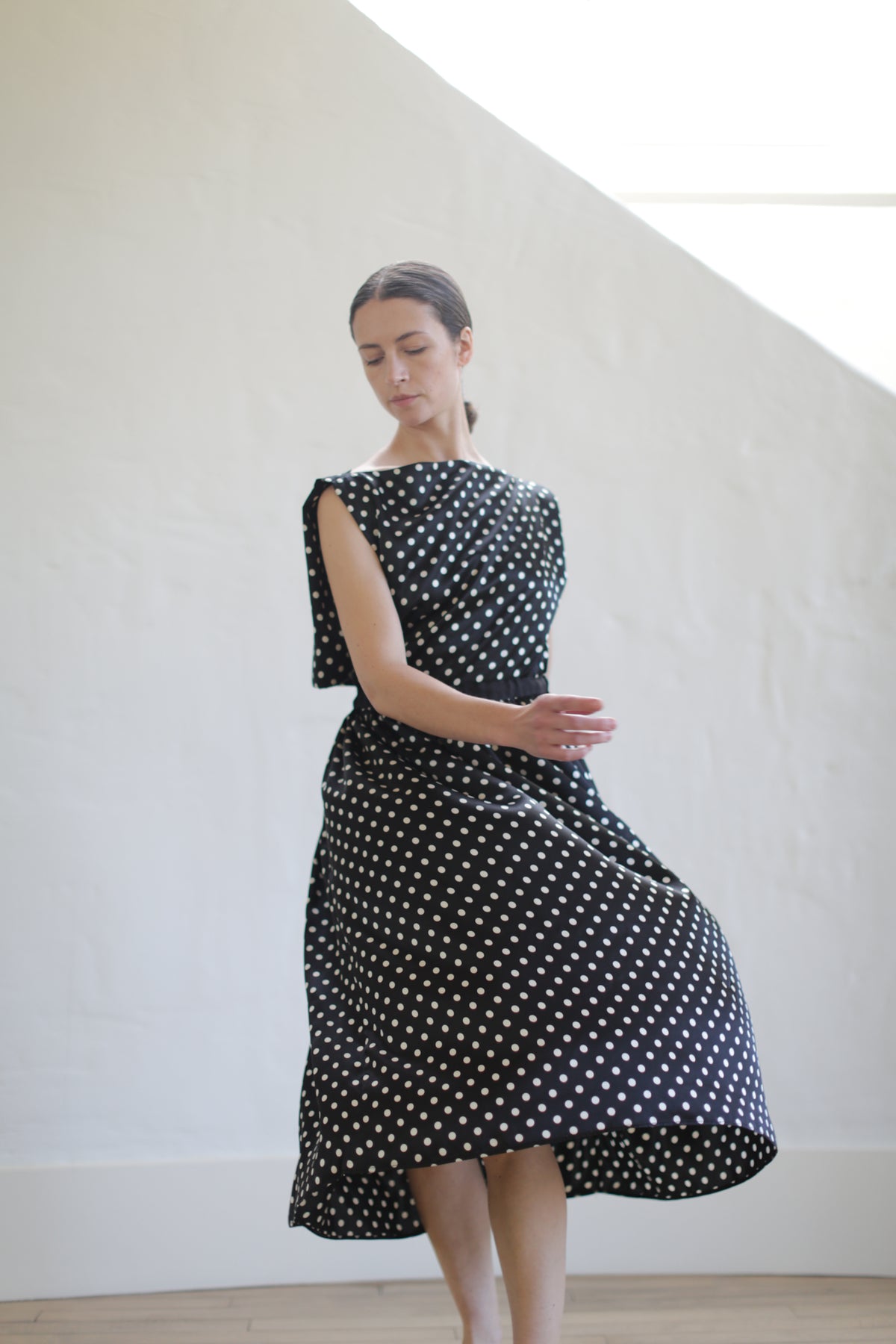 A woman in a sleeveless Magnolia Dress | B/W Polkadot stands gracefully with her eyes closed. She is indoors, against a white wall, and the handmade dress flares as she slightly lifts one leg while her arms are gently extended.