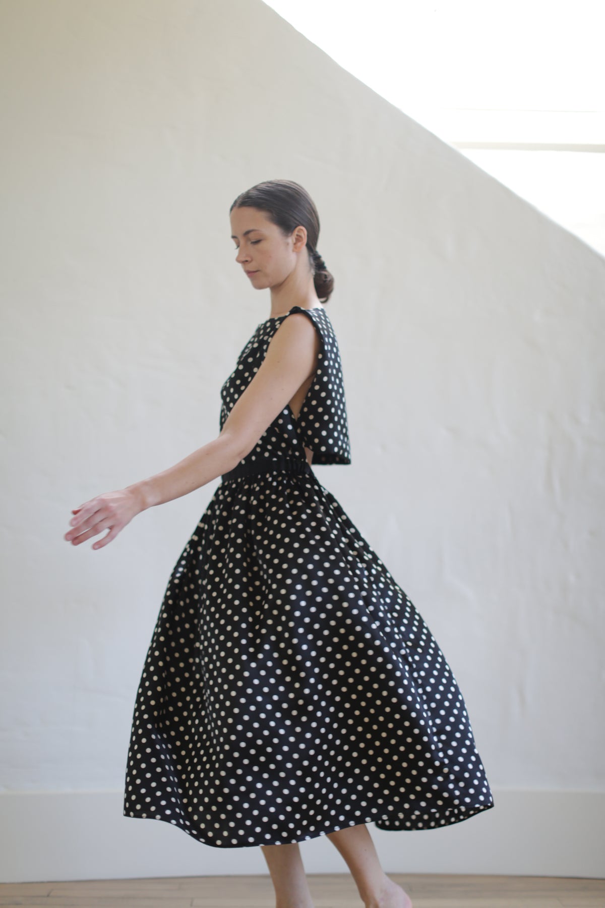 A woman wearing a Magnolia Dress | B/W Polkadot with an open back twirls gracefully in a minimalistic room with a light-colored wall. Her hair is tied back in a ponytail, and she has a serene expression on her face. Her dress flares out as she spins.