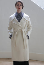 Load image into Gallery viewer, A person stands indoors against a light-colored wall, wearing a Malvern Doubleface Cashmere - Undyed coat with a tie belt and a gray collared shirt underneath. The individual has dark hair pulled back, and hands are in the coat pockets. The background includes a wooden floor.