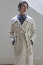 Load image into Gallery viewer, A woman with dark hair styled in a bun is standing against a light background. She is wearing a long, light-colored, double-breasted wrap coat over a collared shirt. The fully lined Malvern Doubleface Cashmere - Undyed coat features a belted waist. She has her hands in her coat pockets and is gazing slightly to the side with a neutral expression.